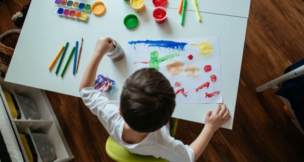 5 year old boy cleaning brush with his left hand from paint in jar with water. Over head view of child with paintbrush and colorful paints on white table at home.